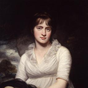 Photograph of a painting of Amelia Opie by her husband, John Opie, 1798. She is depicted from the waist up, against a background that is mainly dark with a landscape scene visible in the distance. She wears a simple white dress with a V-shaped neckline that is trimmed with white ruffles. Her brown hair is pulled back, with bangs, and a braid across the top, acting as a headband. She holds a straw hat wrapped in gauzy black fabric in her lap. National Portrait Gallery.