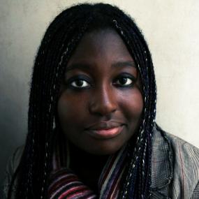 Colour photo portrait of Helen Oyeyemi in London, 2005. In this close-up image, she looks directly at the camera. Her hair is in small braids
            with pale green and blue threads woven through and she is wearing a striped scarf and a beige plaid blazer. 