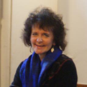 Slightly blurry, half-length photograph of Ruth Padel in Crete, smiling with her arms crossed, wearing a dark jacket. a bright blue scarf, and gold earrings.  Her hair is shoulder-length, curly, and rather wild. In front is a colourful length of woven fabric.