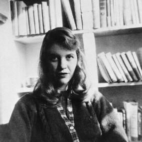Black-and-white photo portrait of Sylvia Plath, looking directly at the camera and seated in front of bookshelves. 