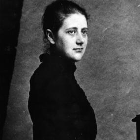 Black-and-white photo portrait of Beatrix Potter taken at Birnamm Scotland in 1892. Standing and looking off-camera, she holds a rose in her
            left hand.