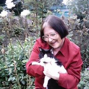 Colour photo of Sheenagh Pugh in a garden, kneeling with a bed of white roses behind her, looking down at a black and white cat in her arms. She has straight, dark hair with grey roots, jaw-length, and wears a red shirt, blue trousers, and glasses with large frames.