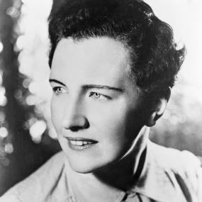 Black-and-white close-up photo of Mary Renault, 1946. Her hair is very short and she is wearing a collared shirt. The background is dappled
            with blurred light.