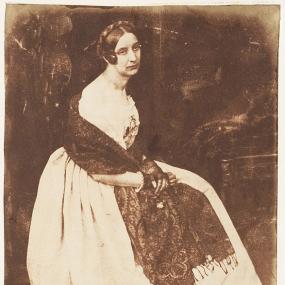 Black and white photograph of Elizabeth Rigby, seated, with her hands clasped in her lap. She is wearing a simple light dress with a long, full skirt, and a dark scarf wrapped around her shoulders. She is wearing small dark gloves with white trim at the wrists, and her dark hair is pulled back, with a braid pulled across the top of her head and another braid wrapped at the side of head.