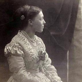 Black and white photograph of Anne Thackeray Ritchie, seated and staring peacefully off into the distance. She is wearing a white dress with puffed shoulders, long sleeves, details of lace, ruffles, and a flower at the neckline, and a pearl button fastening the waistband. She has a stack of pearl necklaces around her neck, her dark hair is pulled into a bun and fastened with a metal pin, and she holds flowers in her hands.