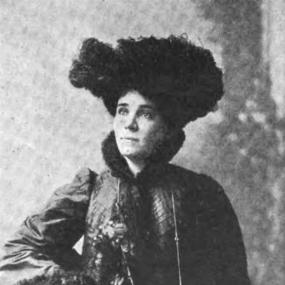 Black and white photograph of Elizabeth Robins, seated, with one arm resting on the arm of her chair and the other in her lap. She is wearing a dark dress with a high collar and long sleeves, both of which are trimmed with fur, a tall, spreading dark hat probably of feathers, and a long silver chain.