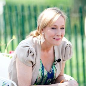 Informal photograph of J. K. Rowling, leaning forward in a white chair in front of a green background, hands clasped on a book in her lap.  She wears a pale cardigan over a colourful dress. Her blonde hair is pulled back and she dangling white teardrop earrings.