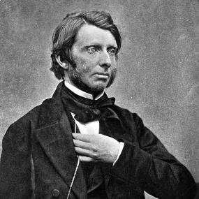 Black and white three-quarter-length image of John Ruskin from a photograph taken at the Working Men's Club. His left hand reaches under the right lapel of his dark jacket, touching a long watch-chain.