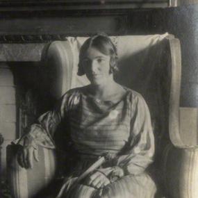 Black and white photograph of Dora Russell, seated in an upholstered armchair in front of a fireplace: the mantlepiece has several ornaments on it. She is wearing a long, horizontally-striped dress with long sleeves and a full pleated skirt. Her dark hair is partly pulled back, with a few curls framing her face.