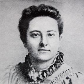 Black and white photo of Olive Schreiner, head and shoulders only. She looks straight at the camera, her hair short around her face and her neckline high, with ruffles.
