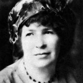 Black and white photograph of Catharine Amy Dawson Scott, shown from the shoulders up. She is wearing a cardigan over a black and white dress, a black beaded necklace, and a puffy hat made of a shiny material, over top of dark curly hair.