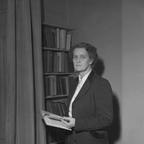 Black-and-white photo of Evelyn Sharp at her home, standing in front of a bookcase and holding a book open in front of her, 16 February
            1951.