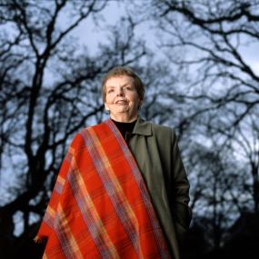 Colour photo of Carolyn Shields taken outside at her home in Victoria, Canada, 1995. Shot from below, she looks off-camera, wears a red tartan
            scarf draped over her right shoulder, and is framed by tree branches overhead in a light blue, near-night sky.