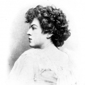 Black and white, head-and-shoulders photograph of Dora Sigerson. She turns away from the camera, her face in profile. She is wearing a simple light dress and has short curly hair.