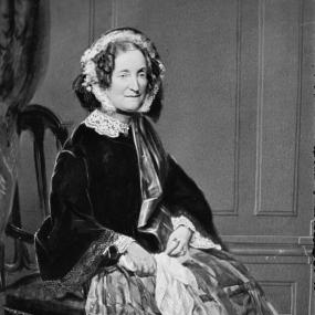 Full-length photograph of Lydia Howard Sigourney by Matthew Brady. She is seated on a wooden chair with wooden panelling behind her, wearing a dark jacket and hood with lace bonnet and collar, and a full, boldly checked skirt. She holds a handkerchief in her hands.