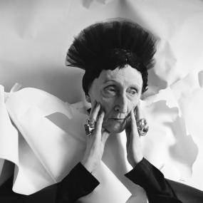 Black and white, head-and-shoulders  photograph of Edith Sitwell by Cecil Beaton, 1962. She wears her black ostrich-feather turban with taffeta standing up on top, a black long-sleeved blouse, and over it an elaborately folded white arrangement like wings. She rests her head between her hands, which are adorned with large, heavy rings.