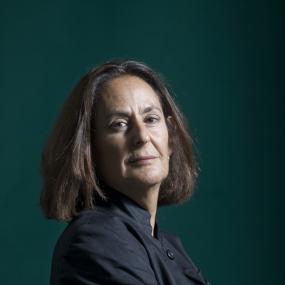 Colour photo portrait of Gillian Slovo at the Edinburgh International Book Festival, August 2009. Looking at the camera, she is wearing a navy
            shacket with her arms crosses in front of her. The background is dark teal.