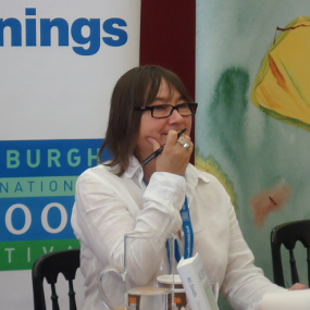 Photograph of Ali Smith, seated at a table, with books, a water jug, and a cup in front of her. She appears to be at a book signing, and she is holding a pen in one hand, which is resting against her chin. She is wearing a white collared shirt dark rectangular-framed glasses, and her brown is shoulder-length with bangs.