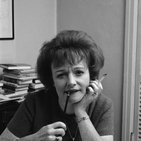 Black-and-white photo of Muriel Spark, 1965. Sitting at a desk, she looks directly at the camera, leaning on her papers with a cigarette in
            her left hand and a pencil in her mouth. 