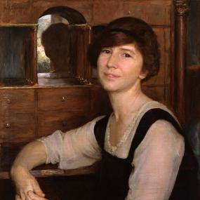 Photograph of a head-and-shoulders painting of Freya Stark by Herbert Arnould Olivier, 1923. She sits in front of a wooden cabinet, with many drawers and an inset mirror, her arm resting on its shelf. She is wearing a simple black tunic over a sheer white blouse, and she has jaw-length brown hair tied with a black ribbon. National Portrait Gallery.