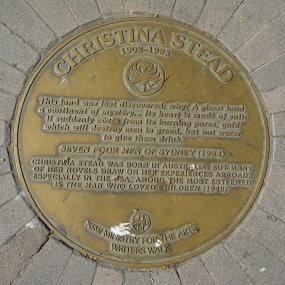 Photograph of a gold-coloured memorial plaque to Christina Stead in the Writers Walk, Sydney (near the Opera House), created in 1991 by the New South Wales Ministry for the Arts. The plaque is set against a grey brick wall, and headed with Stead's name and dates. Below, reading downwards, come a circular emblem of the NSW state flower, telopea speciosissima, a quotation from her "Seven Poor Men of Sydney", a brief summary of her works with special mention of "The Man Who Loved Children", an encircled cross,