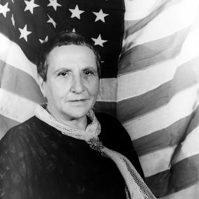 Black and white photo of Gertude Stein taken by Carl Van Vechten in early 1935. She is standing in front of an American flag, wearing a dark jacket and pale scarf.