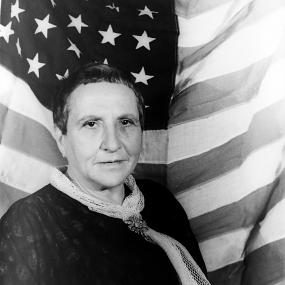 Black and white photo of Gertude Stein taken by Carl Van Vechten in early 1935. She is standing in front of an American flag, wearing a dark jacket and pale scarf.