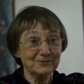 Photograph of Anne Stevenson, shown from the shoulders up, wearing  a  collared shirt, made of multicoloured velvet fabric that blends dark blue, red, and green. She is wearing wire rimmed glasses and a necklace made of copper-coloured metal, and large red stones. Her hair is jaw length and grey.