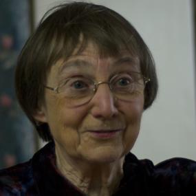 Photograph of Anne Stevenson, shown from the shoulders up, wearing  a  collared shirt, made of multicoloured velvet fabric that blends dark blue, red, and green. She is wearing wire rimmed glasses and a necklace made of copper-coloured metal, and large red stones. Her hair is jaw length and grey.