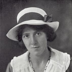 Black and white, head-and-shoulders photograph of Marie Stopes, c. 1918. She has short curly hair, and wears a dress with dark sleeves and broad white collar, a necklace of large beads, and a white Panama-style hat with a dark bow.
