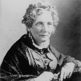 Black and white, head and shoulders photograph of Harriet Beecher Stowe. She is seated, wearing a dark dress with long, wide sleeves, buttons up the front, and a high, ruffled collar. She holds a pair of small spectacles in her hands and her fair, short, curly hair looks unbrushed.
