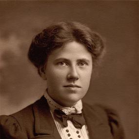 Black and white, head-and-shoulders photograph of Ray Strachey wearing a dark jacket over a white dress shirt with bow tie and two vertical rows of dark buttons. Her dark hair is piled behind her head.