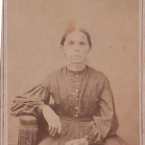 Black and white photograph of Hesba Stretton, seated, with her arm resting on the ornately tasseled arm of her chair. She is plainly dressed in a floor-length dark dress, with buttons up the front and long puff sleeves. Her dark hair is parted in the centre and pulled back tightly; her gaze is straight ahead.