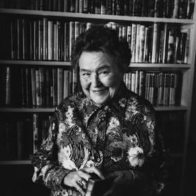 Black-and-white photo of Noel Streatfeild, November 1972. Smiling and looking directly at the camera, she is wearing a floral top, standing
            against a bookshelf, and holding copy of her book Ballet Shoes.
