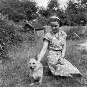 Black-and-white photo of Jan Struther, 28 June 1946. Wearing a patterned dress, she is sitting on the grass and looking down at a small,
            leashed dog beside her.