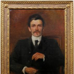 Photograph of oil painting of John Millington Synge, with a gold frame. Synge is wearing a black suit and vest, black tie, and white collared shirt, and he is looking directly at the viewer. His hands appear to be adjusting his suit.