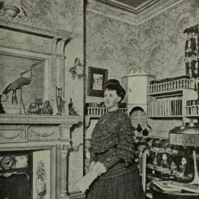 Black and white full-length photograph of Katherine Cecil Thurston standing beside a fire burning in an Adam-style fireplace in a stately home. Her setting is cluttered: the wallpaper is patterned; the fireplace has painted panels, a pediment, and a mantelpiece with large ornaments in front of a mirror; the bookcases have little balustrades and a niche for more ornaments. A black cat sits on the mat. Thurston wears a dark floor-length dress with long sleeves, ruffles, and lace trim. Her dark hair is up in a