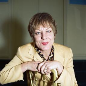 Colour photo of Sue Townsend, 27 October 1992. Looking directly at the camera, she is seated on a gold painted chair and wearing a light
            yellow blazer and skirt over a cheetah-print shirt. 
