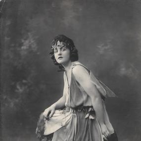 Black and white photograph of P. L. Travers playing the part of Titania in Shakespeare's "Midsummer Night's Dream", c. 1924. She perches on a log wearing a sleeveless tunic, with vine-leaves in her unruly curls, and flowers scattered around.