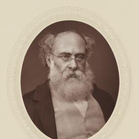 Photograph of woodbury type print depicting Anthony Trollope from the chest up.  He has curly white/grey hair and beard, and is wearing glasses and a suit. His gaze looks slightly to the right, and his brow appears furrowed.