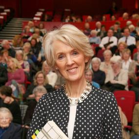 Colour photo of Joanna Trollope standing in a crowded auditorium holding one of her books. She wears a black jacket with white polka-dots over a white shirt, and a chunky metal and pearl necklace. Her blonde hair is chin-length.