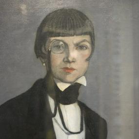 Colour photo of Una Troubridge from the waist up, wearing a black suit-jacket, white high-necked shirt, cravat, and red lipstick. Her brown hair is cropped, but her fringe is quite long. She has a monocle in one eye.