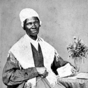 Black and white photo of Sojourner Truth, wearing wire spectacles, a white cap,  long, dark, long-sleeved dress, and a white shawl. She sits beside a small table with a vase of flowers on it, holding a half-done piece of knitting. She regularly used small, carte-de-visite photos to publicize her cause, under the slogan: “I Sell the Shadow to Support the Substance.”