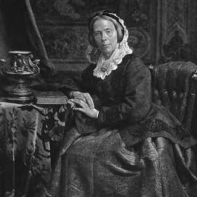 Black and white photograph of Charlotte Maria Tucker by William Notman, Toronto, 1875. She sits in an upholstered chair beside a small table draped with a tablecloth and bearing a book and an ornate Greek-motif jar (probably photographer's props). She wears a long, dark, broad-skirted dress and a simple dark buttoned jacket with white lace collar apparently joined to white lace around a dark cap. Her hair is arranged to shoulder length.
