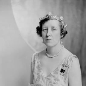 Black and white, half-length photograph of Susan Tweedsmuir, seated, in formal evening dress: a pale sleeveless gown with V neck and two medals pinned  to it, white elbow-length gloves, a white ostrich-feather fan, tiara, necklace, and drop pearl earrings. Her hair is short, curled at the sides.