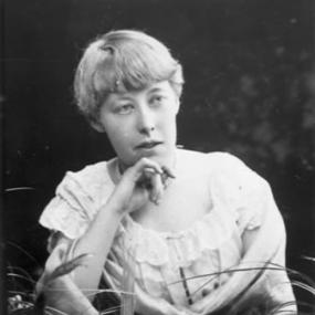 Black and white half-length photograph of a young Katharine Tynan, wearing a white blouse with frilled neckline and short sleeves. Her light hair is short, and she has one hand to her chin. Her surroundings are hard to make out: she may be sitting outdoors with bunches of grasses.