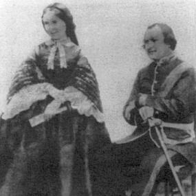 Black and white photograph of Harriet Tytler, standing next to her seated husband, Robert Tytler. She is wearing a long dark dress with a full skirt and wide sleeves trimmed with white lace, with a white bow around her neckline, and holding a fan. Her husband is in British army uniform.
