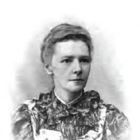 Black and white photograph of Ethel Lilian Voynich, shown from the shoulders up. She is wearing a white smock over a dark dress with long sleeves, and a high, frilled neckline, and her hair is pulled back. Below, her signature is reproduced with a salutation: "Faithfully yours E. L. Voynich."