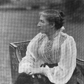 Black and white photograph of Mary Augusta Ward sitting in an upholstered chair, her hair in a bun, hands folded in her lap, and face turned to be seen in profile. She wears a long, full skirt and a gauzy blouse with a high collar, loose sleeves, and patterned panels in front.