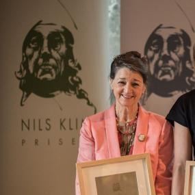 Photograph of Marina Warner receiving the Holberg prize in 2015. She is wearing a patterned shirt, pink blazer, and necklace to match. Her hair is styled up, and she is smiling as she holds her framed prize certificate.
